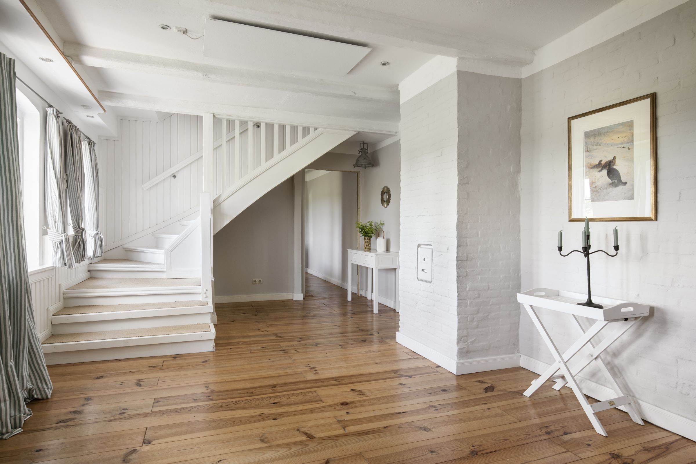 A spacious room with a wooden floor, white walls, a staircase, and a framed picture on the wall.