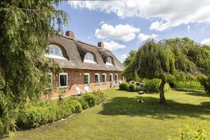 Ein traditionelles Backsteinhaus mit einem strohgedeckten Dach, umgeben von einem gepflegten Garten und Bäumen.