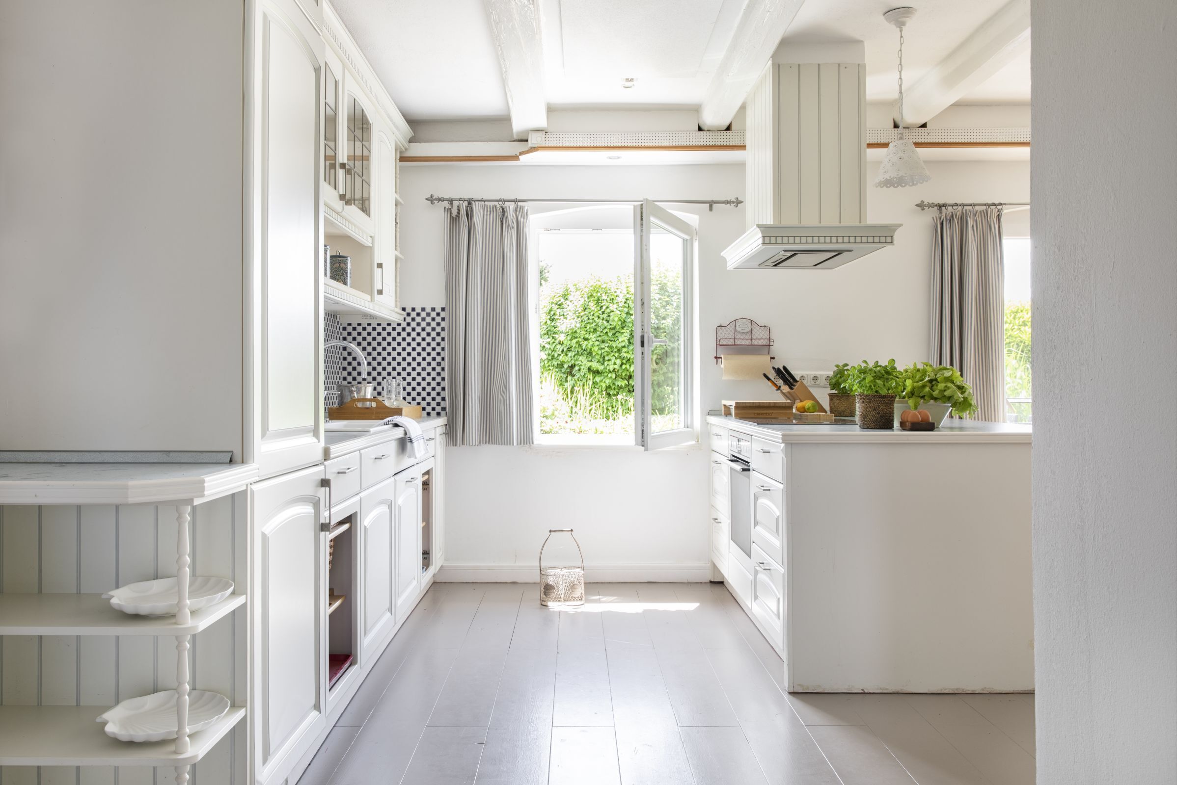 Une cuisine avec des meubles blancs, une fenêtre avec des rideaux et une vue sur la verdure à l’extérieur.