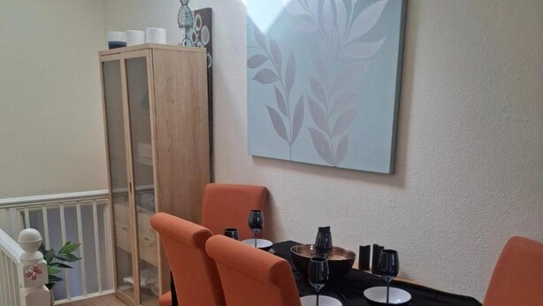 A dining area with a table set for two, chairs, a cabinet, and a wall-mounted artwork. - Barmouth Harbour 2-bed Flat, Sleeps 8 w/ Sea View (Barmouth)