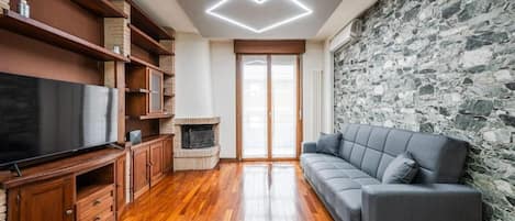 A living room with a fireplace, wooden flooring, a grey sofa, and a stone wall.