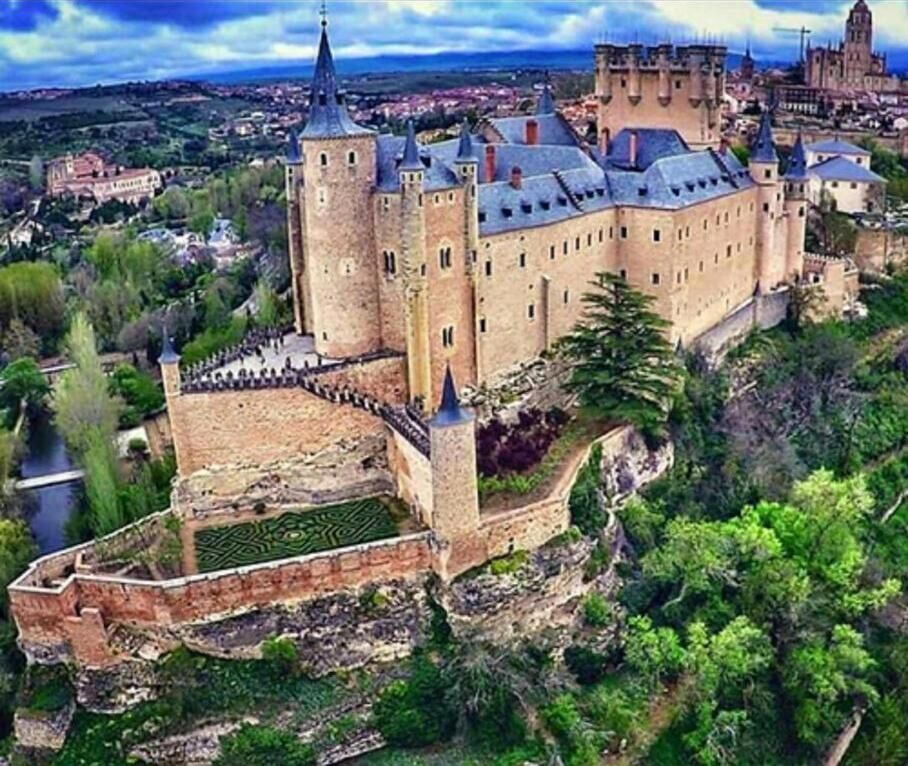 A large castle with multiple towers and a fortified wall, surrounded by greenery and a river.