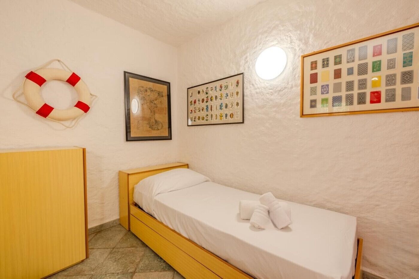 A small, single-bed room with a wooden bed frame, a life preserver on the wall, a framed map, and a collection of patterned tiles.