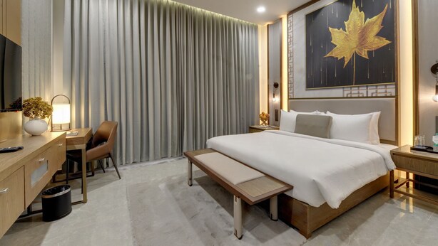 A hotel room with a bed, bedside tables, a desk, a chair, a bench, a TV, and a large wall decoration. - Desert Inn Beacon Greater Rann of Kutch (Bhuj)