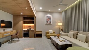 A modern living room with a flat-screen TV, a sofa, a coffee table, and a dining area with a small kitchen setup. - Desert Inn Beacon Greater Rann of Kutch (Bhuj)