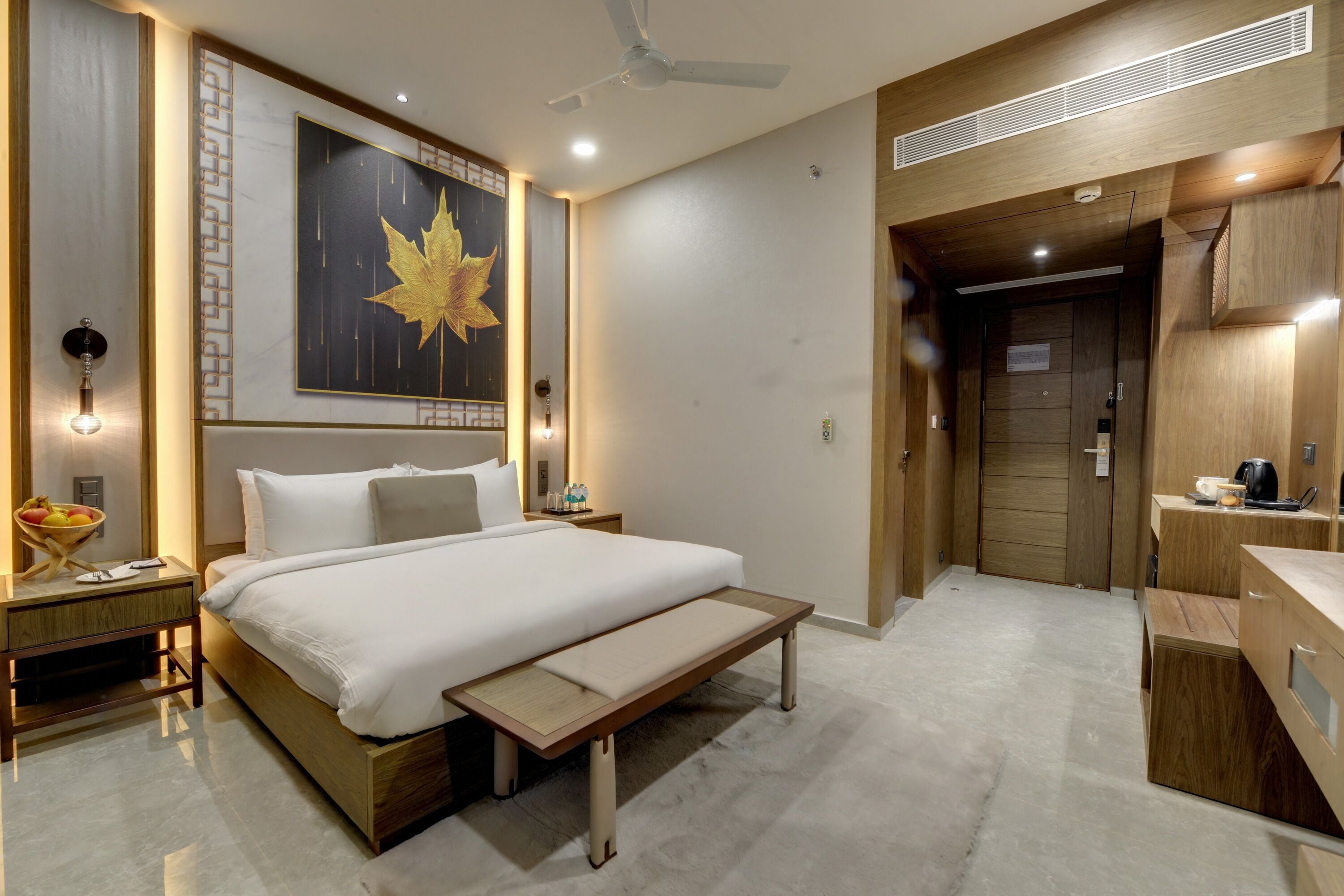 Una habitación de hotel moderna con una cama grande, mesitas de noche, un banco, un frutero y un baño con tocador de madera.