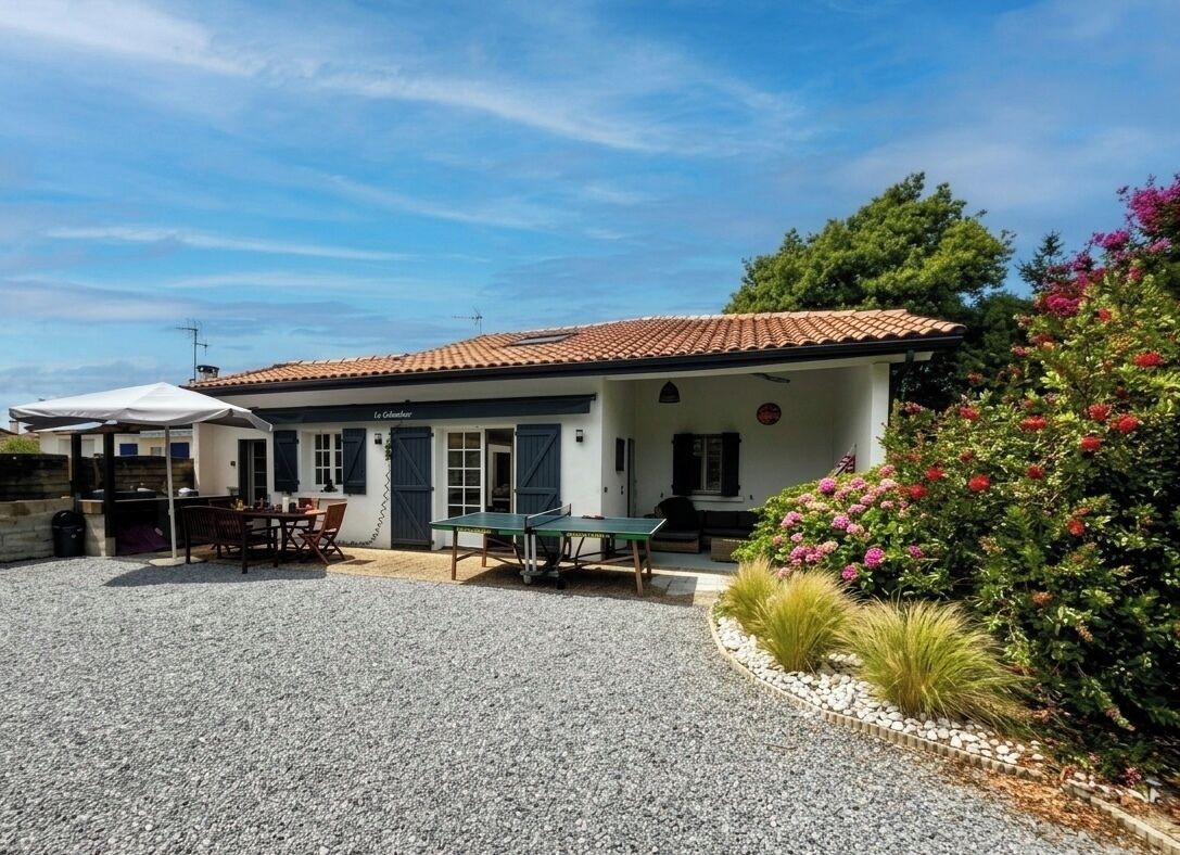 A two-story building with a tiled roof, a covered patio area with a table and chairs, and a garden with flowering plants.