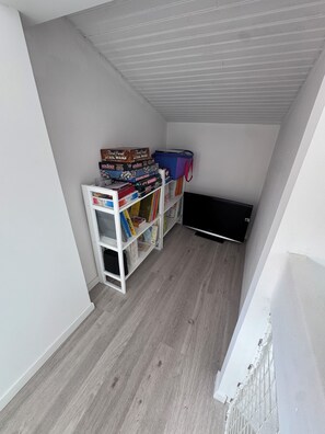 Una habitació amb sostre inclinat, una prestatgeria blanca plena de jocs de taula diversos i un registre negre a la paret.