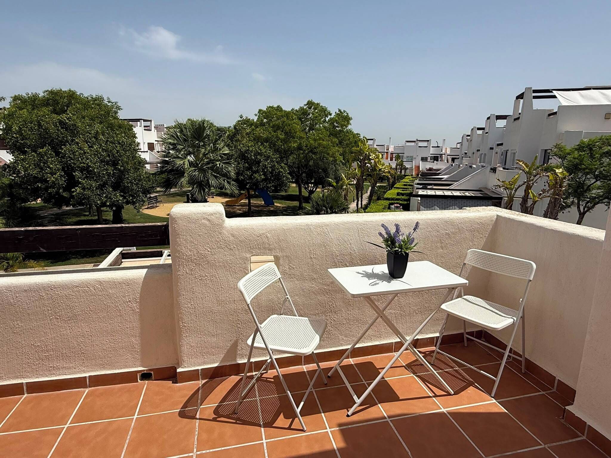 Une terrasse avec une table et deux chaises, un pot de fleurs et une vue sur un quartier résidentiel avec des arbres et des immeubles.