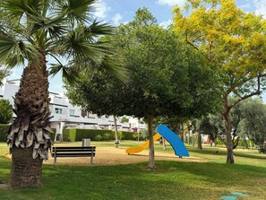 A park with a playground, a palm tree, and a building in the background. - Alhama County - Apartment 186 N3 (MC)