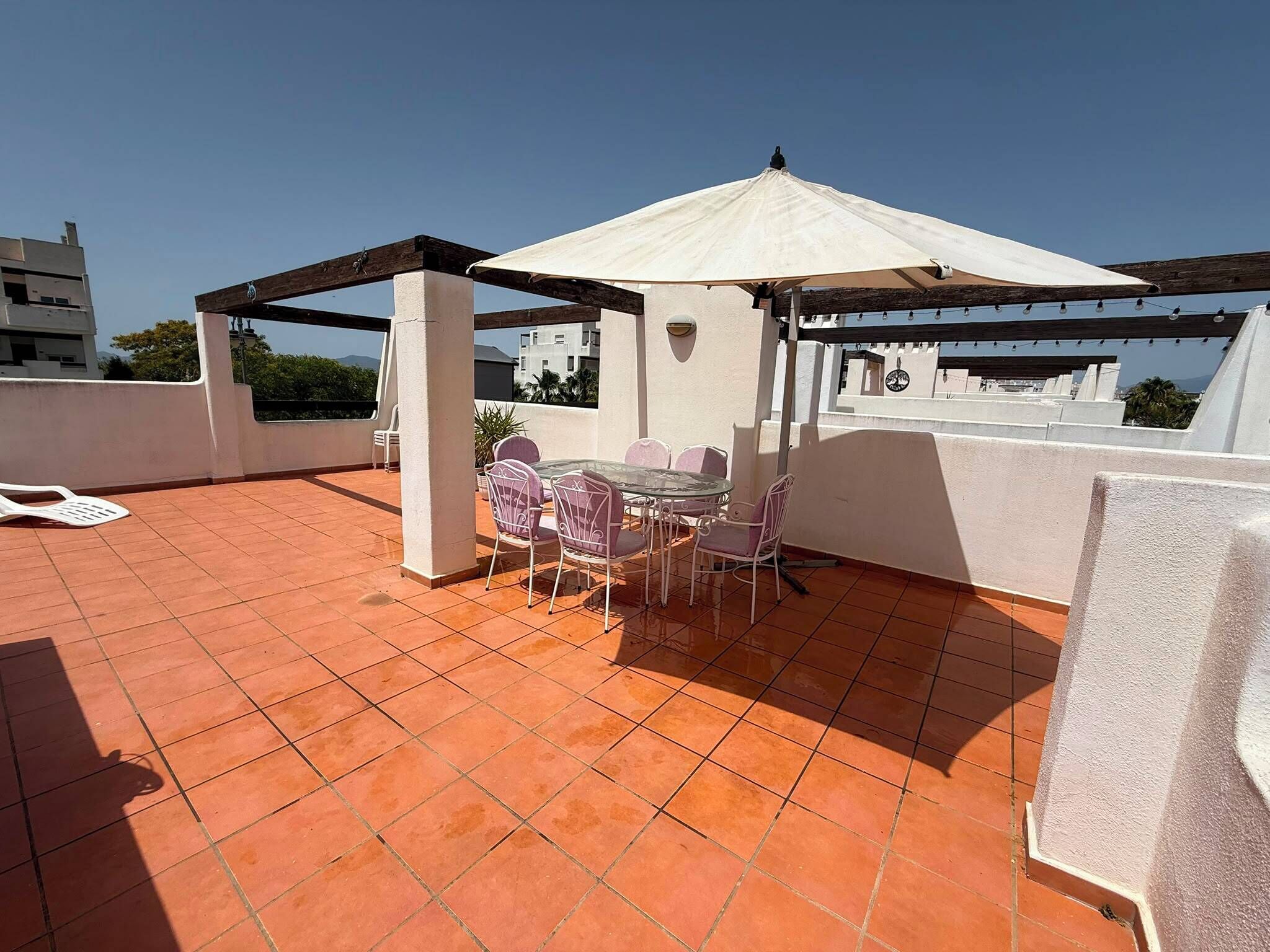 En tagterrasse med et bord og stole under et pergola, omgivet af hvide mure og en klar himmel.