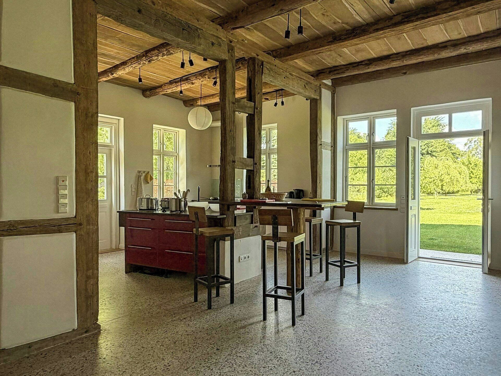 A spacious kitchen with wooden beams, a red cabinet, and a view of a garden through large windows.