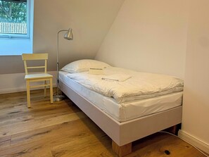 A single bed with a white mattress and pillows, a wooden chair, a floor lamp, and a window with a view of trees.