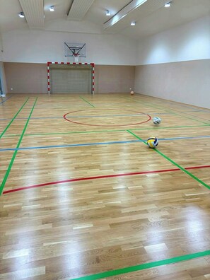 An indoor sports hall with a basketball court, a volleyball net, and two volleyballs.