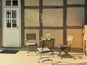 A patio area with a white door, two metal chairs, a small round table with a bowl of fruit and a vase, and a brick wall.