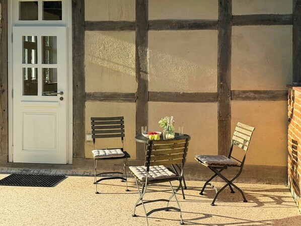 A patio area with a white door, two metal chairs, a small round table with a bowl of fruit and a vase, and a brick wall.