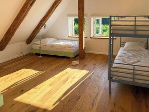 A room with a bunk bed, a single bed, wooden flooring, a window, and a lamp.