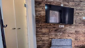 Ein Zimmer mit Fernseher an einer Holzwand, einem grauen Sofa und einer Tür mit Kleiderhaken.
