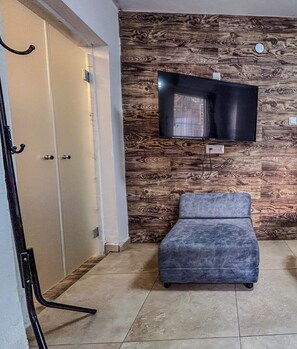 A room with a television mounted on a wooden wall, a grey sofa, and a door with a coat rack.