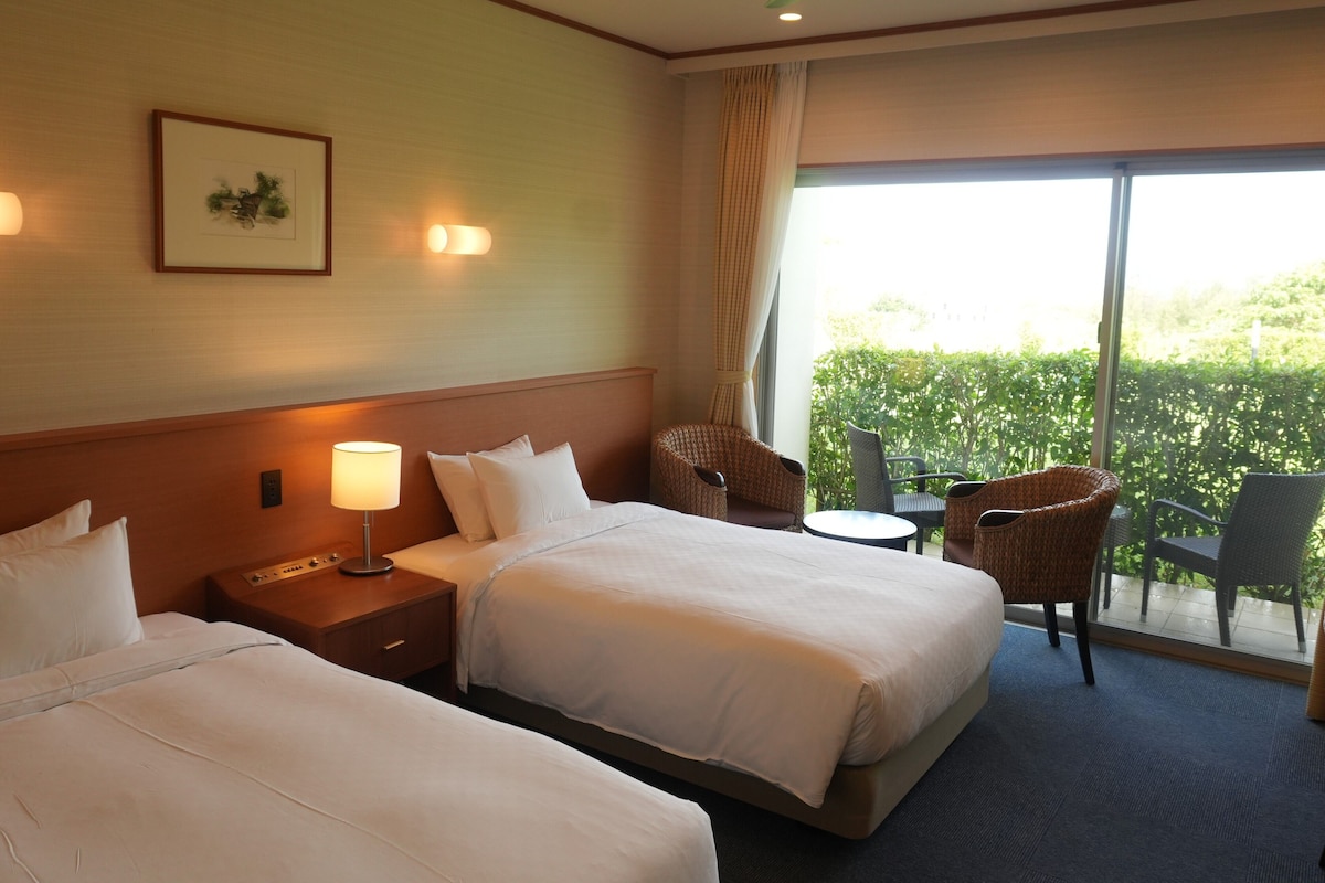 A hotel room with two beds, a ceiling fan, a small table, and a view of greenery outside.