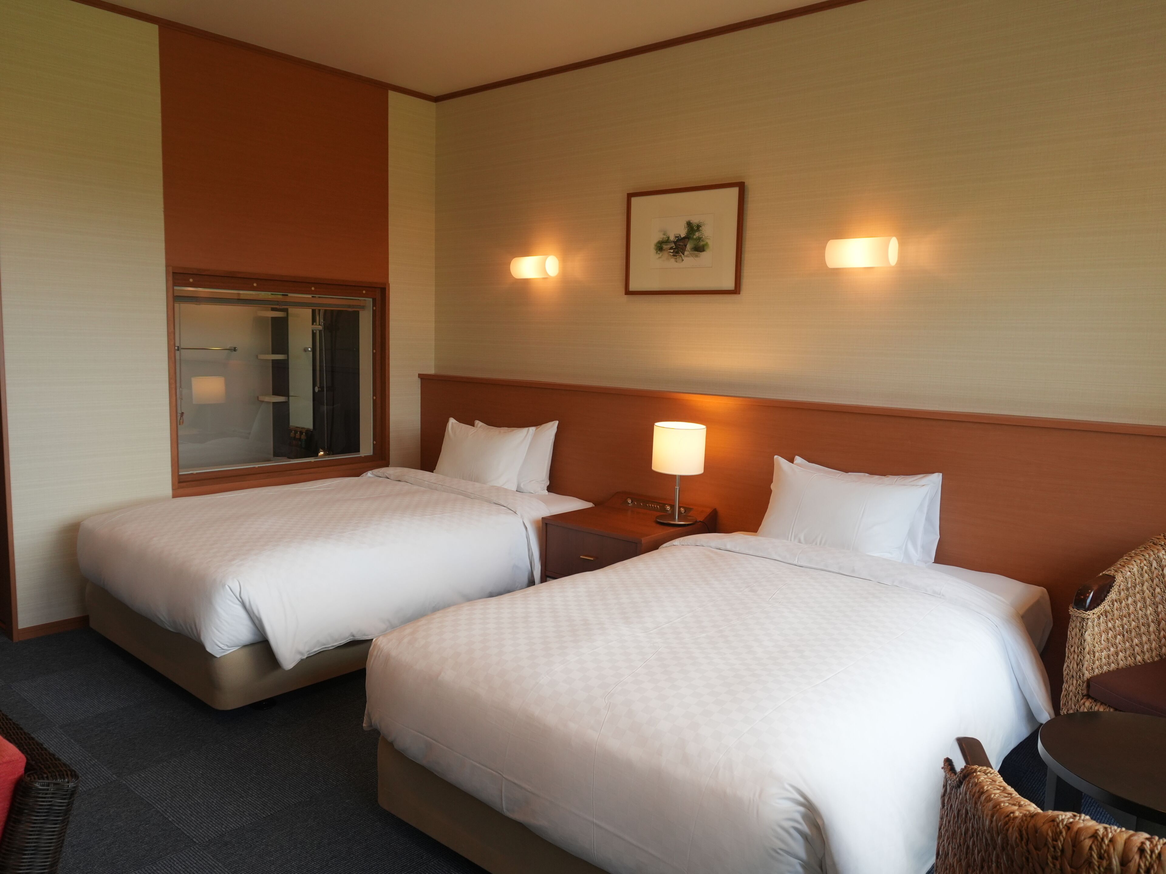 Habitación de hotel con dos camas, una mesita de noche con lámpara, una pequeña mesa y una silla de mimbre.