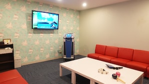 Una habitación con una televisión montada en la pared, un sofá rojo, una mesa blanca con un control remoto, una consola de videojuegos y un papel tapiz estampado.