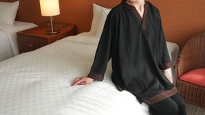 A person standing by a bed with a white bedspread, a wooden nightstand with a lamp, and a wicker chair with a red cushion. - SAKURA RESORT HOTEL ISHIGAKI (Ishigaki)