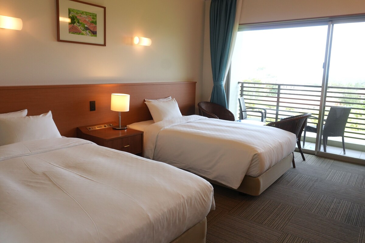 A hotel room with two beds, a nightstand, a lamp, a chair, and a balcony with a railing.