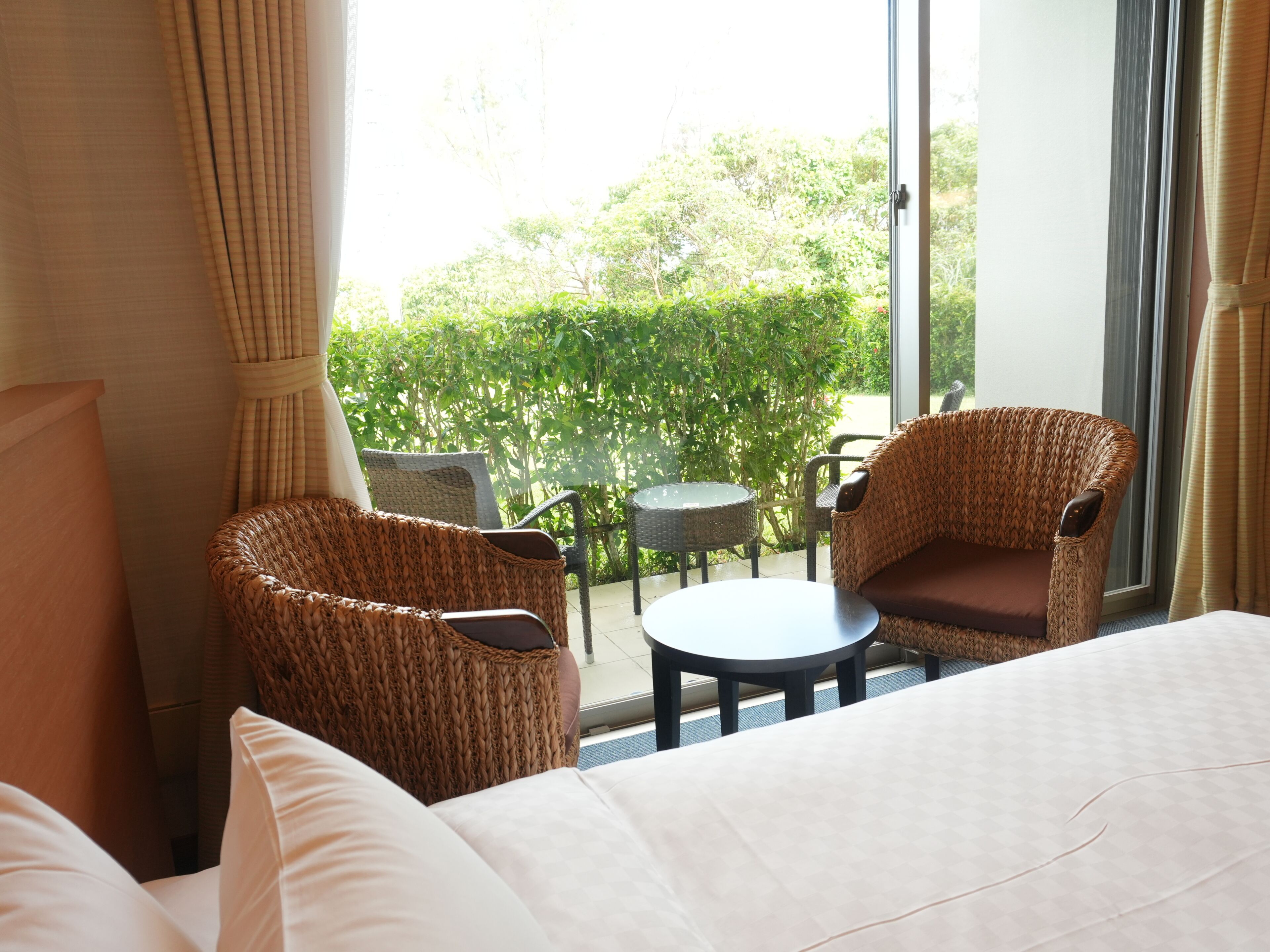 Quarto de hotel com cama, duas cadeiras de vime, uma mesa redonda pequena e vista para o verde através de uma grande janela.