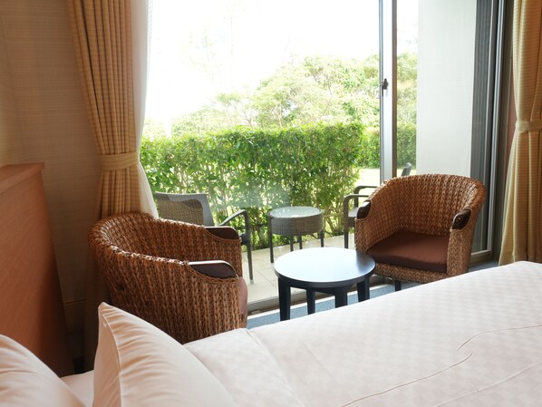 A hotel room with a bed, two wicker chairs, a small round table, and a view of greenery through a large window.