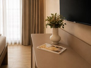 A hotel room with a bed, a bedside table with a vase and a magazine, and a TV mounted on the wall. - Hotel AMANO München Stachus (Munich)