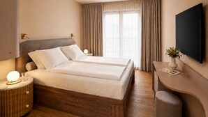 A hotel room with a double bed, bedside tables, a TV, and a desk with a chair. - Hotel AMANO München Stachus (Munich)