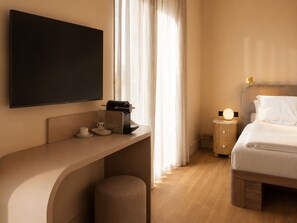 A hotel room with a flat-screen TV mounted on the wall, a bedside table with a lamp, a bed with white bedding, and a window with curtains. - Hotel AMANO München Stachus (Munich)