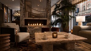 A modern lounge area with a marble coffee table, seating, a fireplace, and a view of the city. - Hotel AMANO München Stachus (Munich)