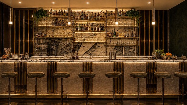 A bar with multiple stools and shelves stocked with bottles and glasses. - Hotel AMANO München Stachus (Munich)