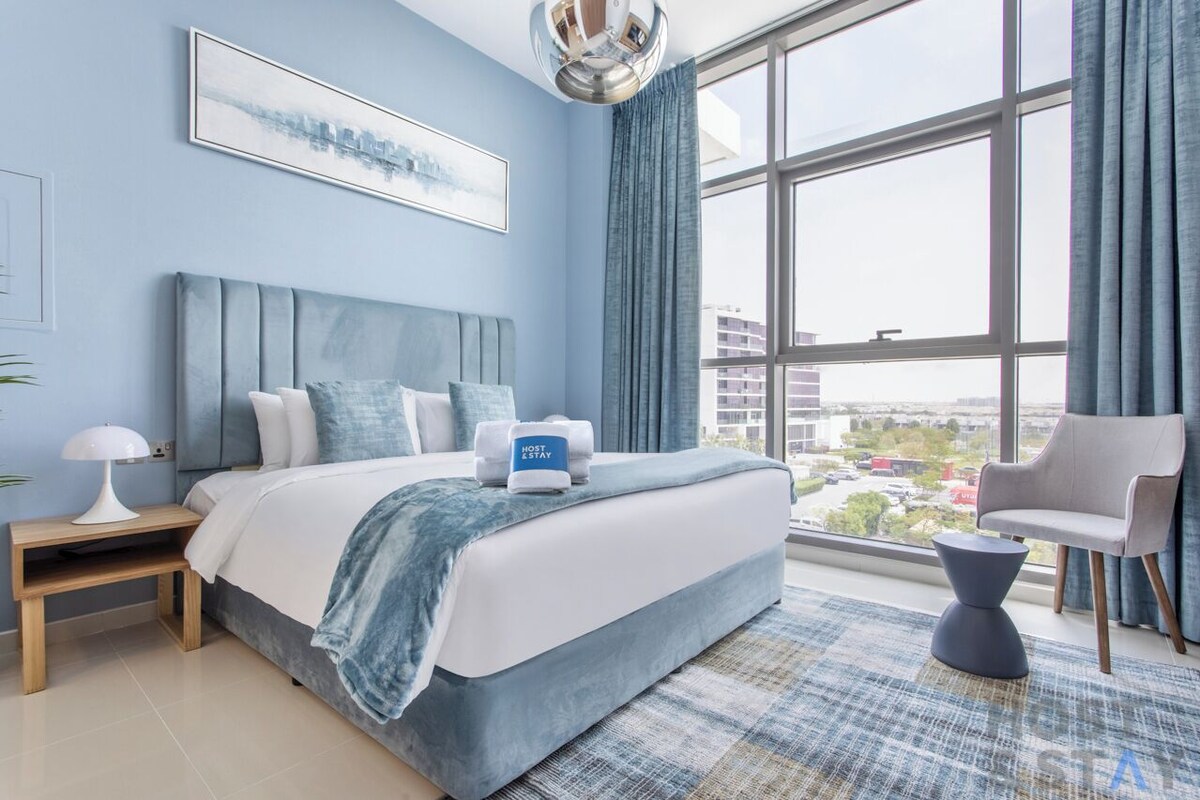 Ein modernes Hotelzimmer mit einem großen Bett, einem Nachttisch, einer Lampe, einem Sessel und Blick auf die Stadt durch große Fenster.
