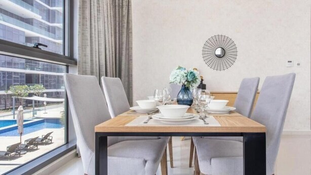 A dining area with a table set for four, a vase of flowers, and a view of a swimming pool. - Orchid A Damac Hills - Host & Stay (Dubai)
