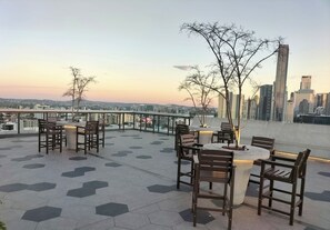 A rooftop terrace with hexagonal tiles, wooden chairs, and tables, overlooking a cityscape with buildings. - Luxe-valley 2BR 2bath Infinity Pool (Brisbane)