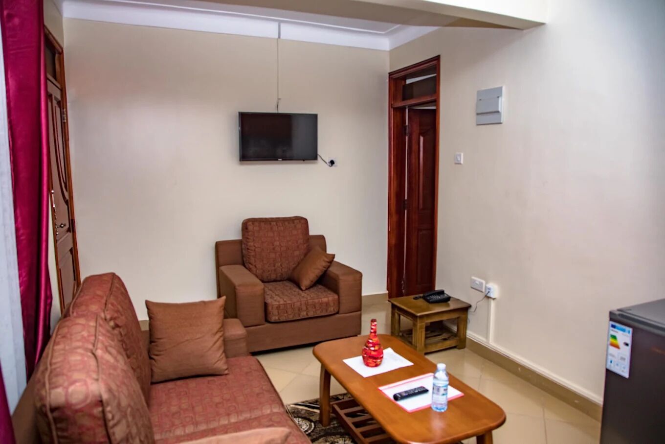 A living room with a sofa, a chair, a TV, a small table with a bottle and remote, and a closed door.