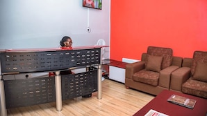A room with a desk, a red wall, a brown sofa, and a television mounted on the wall. - Izera Suites (Kampala)