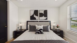 Un dormitorio con cama, dos mesitas de noche, una lámpara de pared, una planta en maceta y una ventana con persianas.