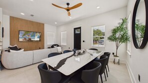 A modern living room with a sofa, a dining table with chairs, a ceiling fan, and a large window with blinds.