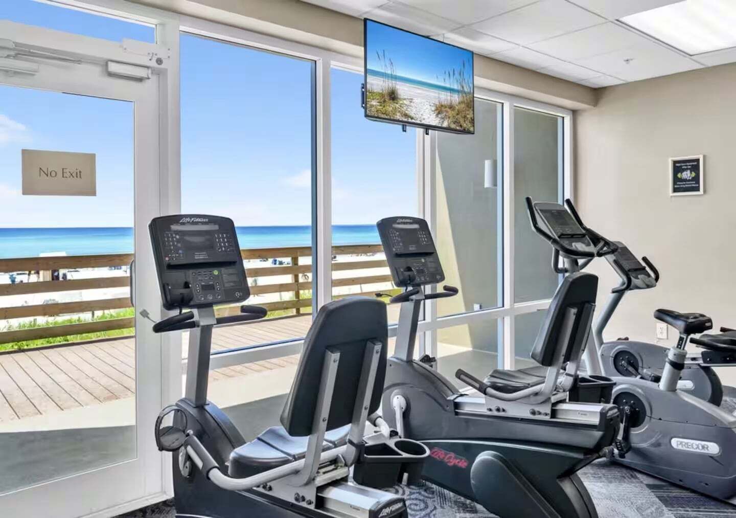 Academia com máquinas de cardio, televisão e vista para a praia.