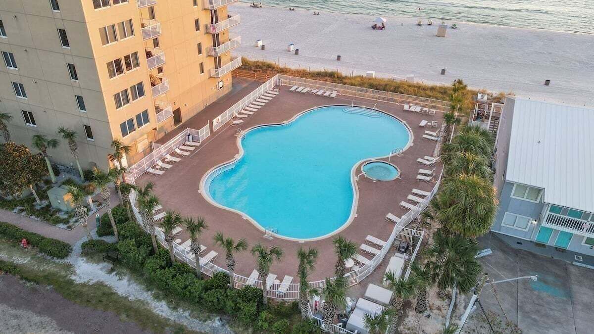 A pool surrounded by lounge chairs, a building with balconies, and a beach in the background.
