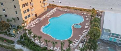 A pool surrounded by lounge chairs, a building with balconies, and a beach in the background.