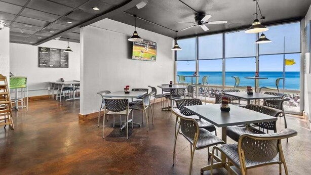 En restaurang med bord och stolar, en takfläkt och ett stort fönster med utsikt över havet.