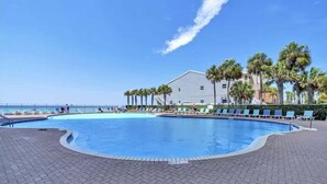 En swimmingpool med solstolar och palmer, en byggnad i bakgrunden och en klar himmel.