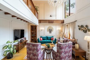 A spacious living area with a high ceiling, a television, a wooden bookshelf, a teal sofa, patterned armchairs, and a coffee table. - Staymaster Shangrila-4BR Private villa  (Marra)