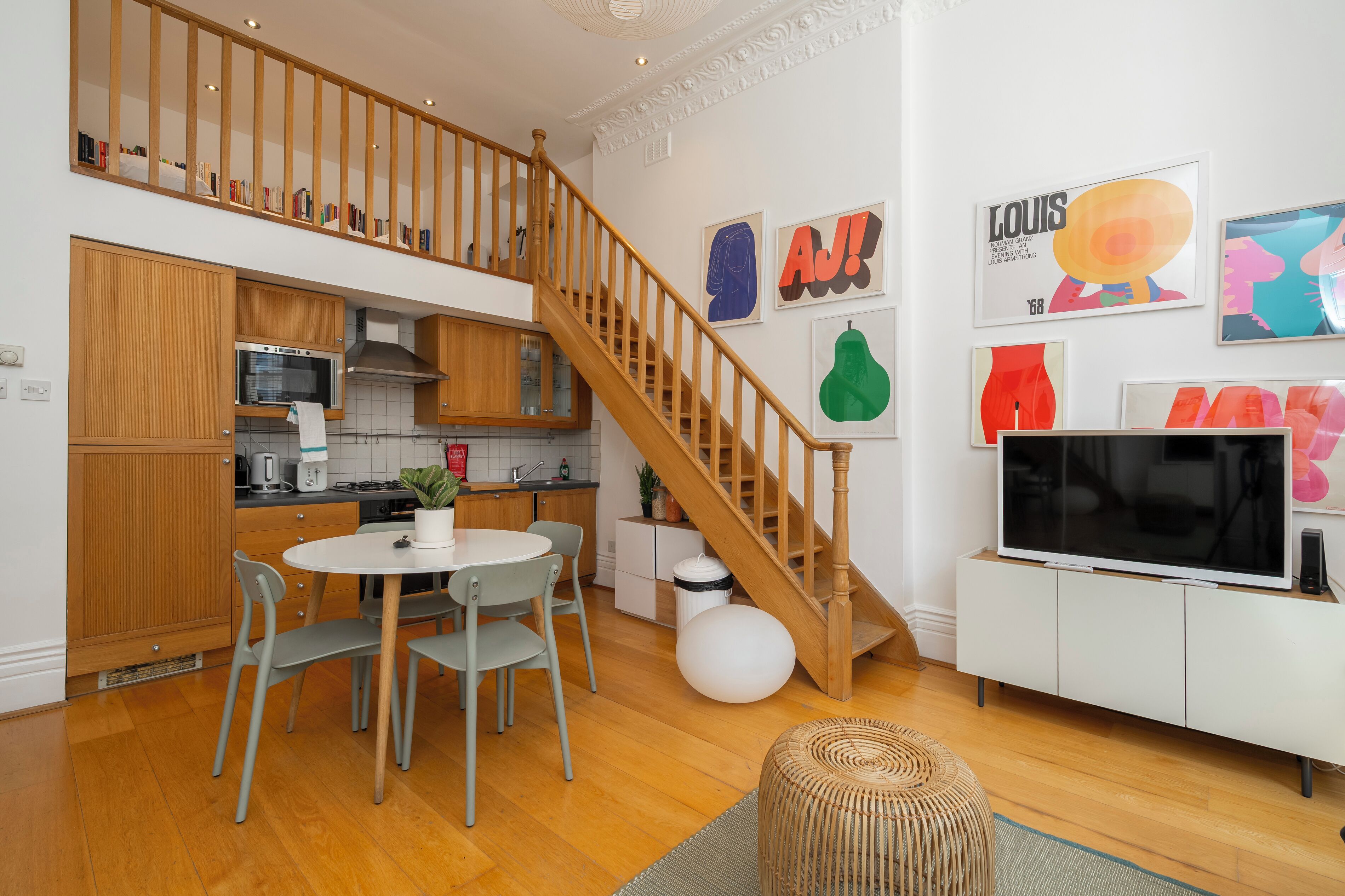 A modern kitchen with wooden cabinets, a dining area with a round table and chairs, a staircase, and a living area with a TV and artwork on the wall.