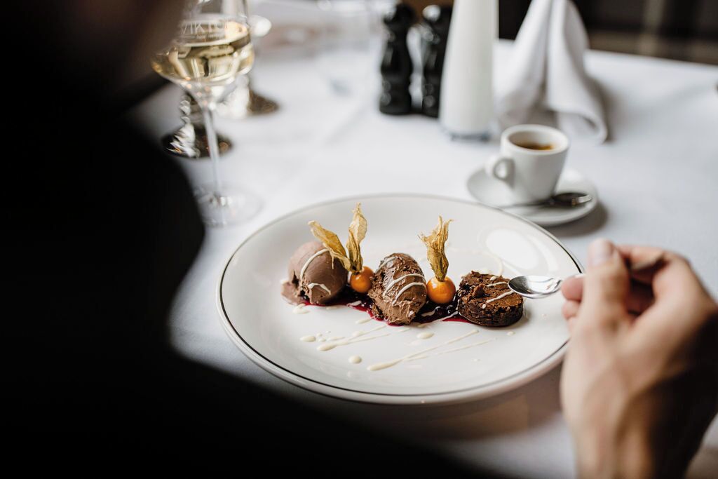 Un plato con helado y frutos rojos sobre una vajilla blanca, una copa de vino blanco y una taza de café en una mesa.
